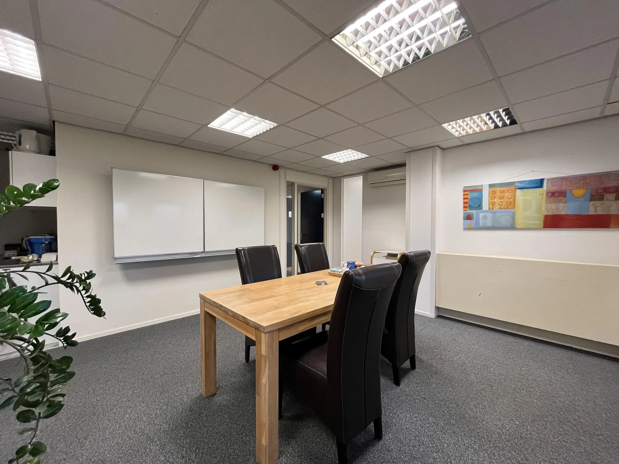 Vergaderruimte in Leidse Rijn met houten tafel, vier zwarte stoelen, whiteboard en kleurrijk kunstwerk aan de muur.